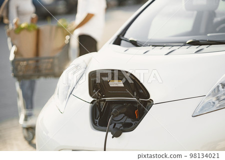 African Arabian couple stands with groceries near electric car. Charging electric car at the electric gas station African Arabian couple stands with groceries near electric car. Charging electric car at the electric gas station 98134021