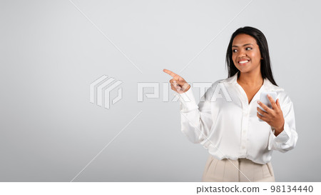 Happy pretty millennial black female in white blouse typing on smartphone, showing finger on free space 98134440
