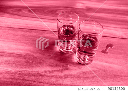 Two shot glasses with whiskey on a wooden table. Image tonned with Viva Magenta color of year 2023. 98134800