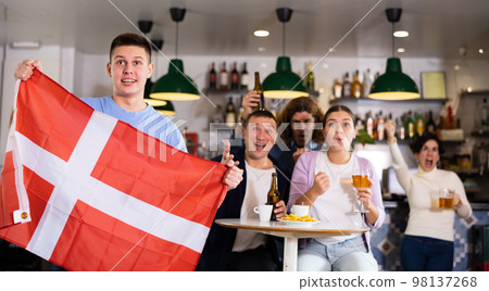Screaming young adult sports fans rooting for favorite team and waving flag of Denmark while watching match together in pub 98137268