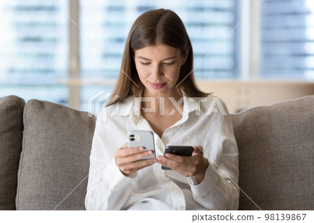 Woman holding two modern smart phones seated on couch 98139867