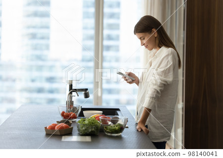 Young woman using smartphone in kitchen while cooking vegetarian salad 98140041