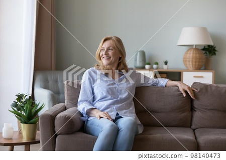 Cheerful attractive mature pensioner woman sitting on home sofa Cheerful attractive mature pensioner woman sitting on home sofa 98140473