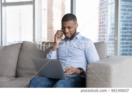 Millennial African man talks on cellphone use laptop at home 98140607