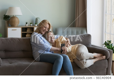 Cheerful mature mom cuddling young adult pretty daughter with smartphone Cheerful mature mom cuddling young adult pretty daughter with smartphone 98140904
