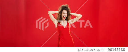Girl just wanna have fun. Crazy adult caucasian woman with curly hairstyle in elegant dress holding hands on head yelling out loud with closed eyes and intense expression over red background Girl just wanna have fun. Crazy adult caucasian woman with curly hairstyle in elegant dress holding hands on head yelling out loud with closed eyes and intense expression over red background 98142688