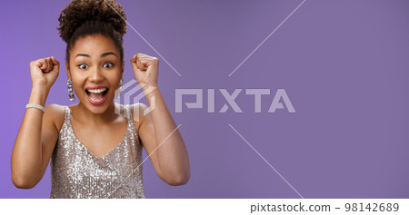 Happy astonished african american woman in silver fashionable dress winning first prize gladly celebrating raising clenched fists dancing triumphing happily having fun, standing blue background 98142689