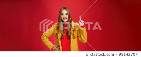 Girl telling first reason use her advice. Cute and friendly energized young redhead woman in yellow coat raising hand showing number one and smiling broadly at camera against red background 98142707
