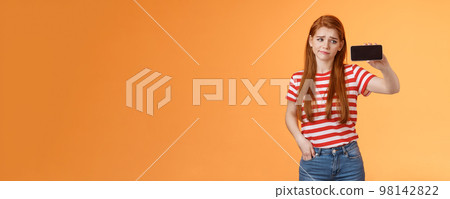 Hesitant disappointed cute redhead woman smirk displeased, hold smartphone horizontal, look device upset, doubtful about blog popularity, sigh uneasy, stare gadget perplexed, show telephone screen Hesitant disappointed cute redhead woman smirk displeased, hold smartphone horizontal, look device upset, doubtful about blog popularity, sigh uneasy, stare gadget perplexed, show telephone screen 98142822