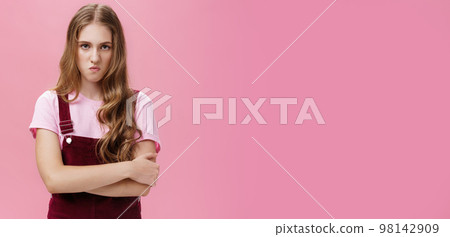 I mad on world. Unhappy hateful young arrogant girl in overalls with wavy natural hairstyle crossing arms against chest in defensive pose lifting upper lip in scorn and disdain, looking with anger 98142909