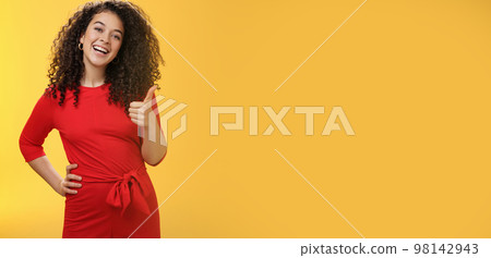 Portrait of happy optimistic young curly-haired woman in red dress laughing joyfully, showing thumbs up in approval and like gesture, delighted with awesome idea, accepting plan over yellow wall Portrait of happy optimistic young curly-haired woman in red dress laughing joyfully, showing thumbs up in approval and like gesture, delighted with awesome idea, accepting plan over yellow wall 98142943