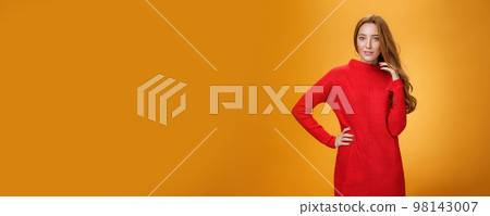 Attractive sensual and romantic redhead woman in red knitted elegant dress holding hand on hip smiling mysteriously as flirting playing with hair gazing at camera over orange background Attractive sensual and romantic redhead woman in red knitted elegant dress holding hand on hip smiling mysteriously as flirting playing with hair gazing at camera over orange background 98143007