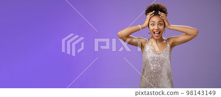 Amazed surprised excited arican-american woman winning lottery cannot believe thrilled hold head screaming amused smiling astonished standing impressed in glittering shiny dress, blue background 98143149