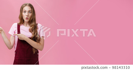 Excited stunned and surprised charming young girl with makeup and curvy hairstyle in trendy corduroy dungarees pointing left folding lips from interest and amazement being curious what happened 98143310