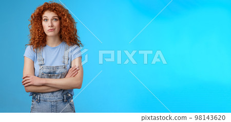Unsure confused redhead curly woman look perplexed uncertain cross arms chest smirking stare camera full disbelief suspicious doubtful someone telling truth standing denim overalls blue background 98143620