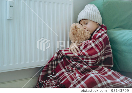 cute little boy wrapped id plaid wearing knit hat sitting by heater hugging teddy bear, cold at home cute little boy wrapped id plaid wearing knit hat sitting by heater hugging teddy bear, cold at home 98143799