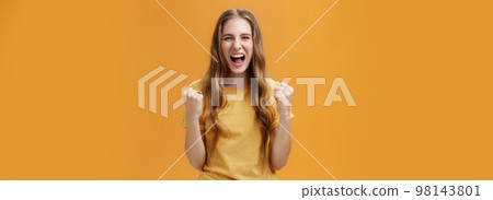 Portrait of happy cheerful lucky young european girl with long natural wavy hair raising clenched fists in cheer and joy yelling yes from triumph feeling glorious moment of win over orange wall 98143801