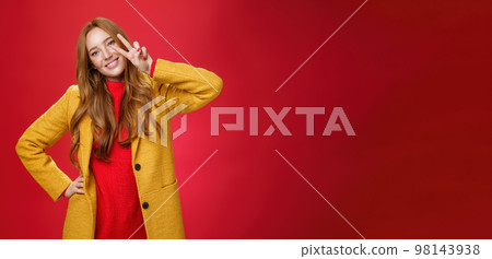 Friendly girl smiling happily, tilting head and showing peace or victory gesture near face, holding hand on hip as posing in stylish elegant yellow coat and sweater against red background Friendly girl smiling happily, tilting head and showing peace or victory gesture near face, holding hand on hip as posing in stylish elegant yellow coat and sweater against red background 98143938