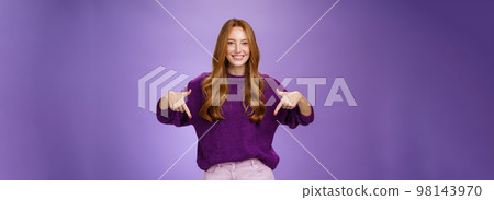 Girl giving recommendation to visit place, pointing down and smiling friendly with delighted and pleasant grin. Portrait of attractive redhead female model in purple warm sweater promoting copy space 98143970