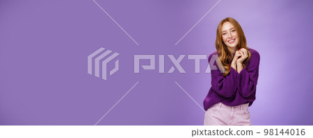 Charming and tender carefree redhead girlfriend with cute freckless holding hands together near shoulder as posing flirty and feminine smiling broadly having positive, happy attitude over purple wall 98144016