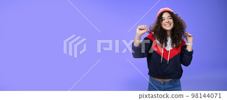 Charming carefree young 20s girl with curly haircut in warm beanie dancing and having fun joyfully over blue wall raising hands relaxed, chill smiling broadly at camera liking perfect chilly weather Charming carefree young 20s girl with curly haircut in warm beanie dancing and having fun joyfully over blue wall raising hands relaxed, chill smiling broadly at camera liking perfect chilly weather 98144071