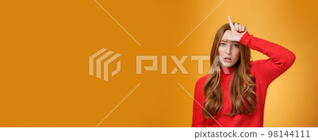 Arrogant and self-satisfied redhead woman humiliating person showing loser sign on forehead mocking and disdain rival standing confident and snobbish over orange background 98144111