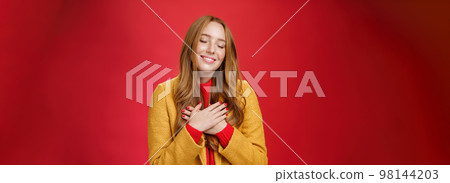 Girl feeling like heart warming as remembering sweet tender memories with closed eyes and silly broad smile holding hands on chest pressing to soul posing nostalgic and touched over red background Girl feeling like heart warming as remembering sweet tender memories with closed eyes and silly broad smile holding hands on chest pressing to soul posing nostalgic and touched over red background 98144203