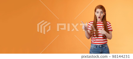 Wow awesome. Impressed excited beautiful redhead woman look astonished explain amazing news, stare camera thrilled talking about advertisement, pointing left upbeat impressed, orange background 98144231