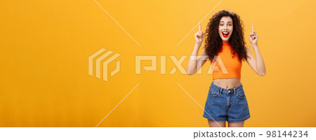 Woman surprising boyfriend with cool sign hanging upstairs pointing up, smiling singing happy b-day standing carefree and joyful over orange background in stylish cropped top and summer denim shorts Woman surprising boyfriend with cool sign hanging upstairs pointing up, smiling singing happy b-day standing carefree and joyful over orange background in stylish cropped top and summer denim shorts 98144234