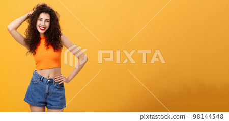 Friendly and stylish modern curly-haired female with red lisptick in denim shorts and cropped top playing with hair smiling flirty and carefree holding hand on waist posing over orange background Friendly and stylish modern curly-haired female with red lisptick in denim shorts and cropped top playing with hair smiling flirty and carefree holding hand on waist posing over orange background 98144548