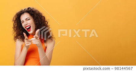Hey what is up. Friendly and outgoing attractive curly-haired female with red lipstick pointing at camera with finger guns as if greeting or hinting friend winking at camera over orange background Hey what is up. Friendly and outgoing attractive curly-haired female with red lipstick pointing at camera with finger guns as if greeting or hinting friend winking at camera over orange background 98144567