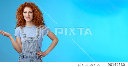 Let me show you cool product. Attractive cheerful confident redhead female model curly hairstyle present customers object hold palm blank blue copy space advertising standing blue background Let me show you cool product. Attractive cheerful confident redhead female model curly hairstyle present customers object hold palm blank blue copy space advertising standing blue background 98144580