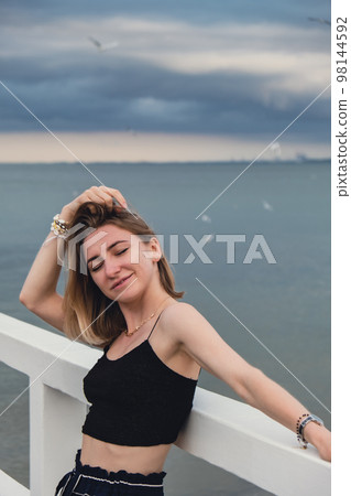 Young woman standing on wooden pier blurred beachside background. Attractive female enjoying the sea shore travel and active lifestyle concept. Springtime. Wellness wellbeing mental health inner peace Young woman standing on wooden pier blurred beachside background. Attractive female enjoying the sea shore travel and active lifestyle concept. Springtime. Wellness wellbeing mental health inner peace 98144592