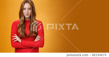Strong and confident good-looking stylish redhead woman with daring gaze and self-assured attitude holding hands crossed over body looking focused at camera over orange background Strong and confident good-looking stylish redhead woman with daring gaze and self-assured attitude holding hands crossed over body looking focused at camera over orange background 98144674