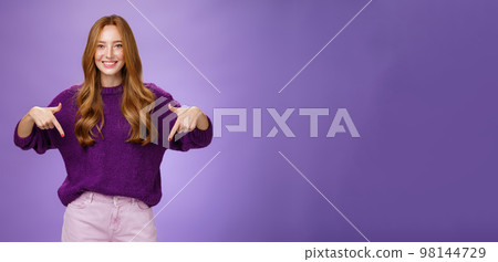 Girl giving recommendation to visit place, pointing down and smiling friendly with delighted and pleasant grin. Portrait of attractive redhead female model in purple warm sweater promoting copy space Girl giving recommendation to visit place, pointing down and smiling friendly with delighted and pleasant grin. Portrait of attractive redhead female model in purple warm sweater promoting copy space 98144729