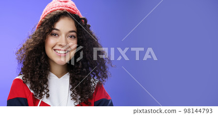 Headshot of tender and cute curly-haired 20s woman in warm beanie and cool sweatshirt smiling broadly enjoying awesome sunny and chilly days outdoors having fun posing over blue background 98144793