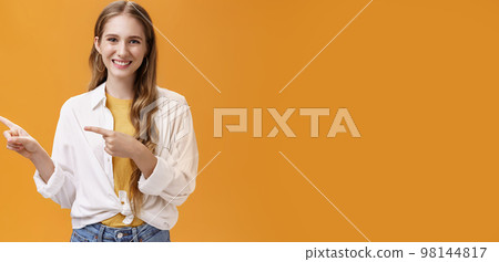 Waist-up shot of friendly pleasant and stylish young teenage female in blouse over t-shirt with cute wavy fair hair pointing left and smiling broadly at camera posing happy against orange background 98144817