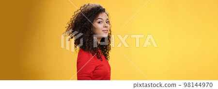 Profile shot of stylish and cool attractive curly-haired european 25s woman in red dress turning at camera and smiling broadly with delighted carefree expression, having fun over yellow background 98144970