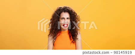 Girl being disgusted with row food sticking out tongue and frowning showing aversion and disgust talking about thing she dislikes with friend standing intense and dissatisfied over orange background 98145919