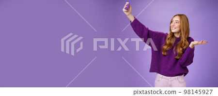 Portrait of cute redhead girl on vacation taking selfies near sightseeing raising hand as if holding builting on palm or showing cool place as taking photograph with pulled arm and smartphone 98145927