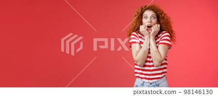 Shocked scared ambushed redhead curly-haired cute girlfriend insecure gasping drop jaw scream frightened stare camera hold hands opened mouth terrified standing stupor red background 98146130