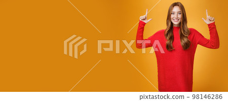 Enthusiastic and excited energized redhead female in red warm sweater pointing up with rised hands and smiling broadly thrilled with awesome promotion, feeling astonished against orange background 98146286