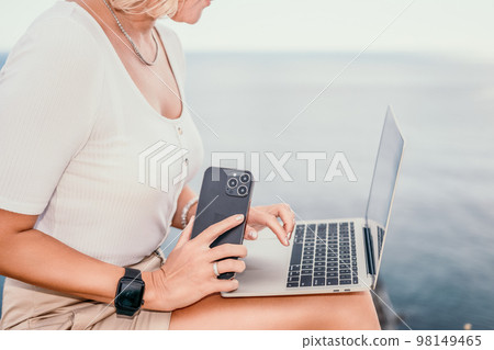 Digital nomad, Business woman working on laptop by the sea. Pretty lady typing on computer by the sea at sunset, makes a business transaction online from a distance. Freelance, remote work on vacation 98149465