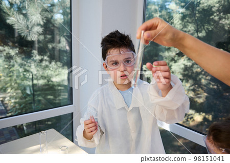 Handsome teenage schoolboy in lab coat and goggles, attentively watching and analyzing the solution in test tube, at chemistry class in school laboratory. Kids and science. Back to school. Education. 98151041