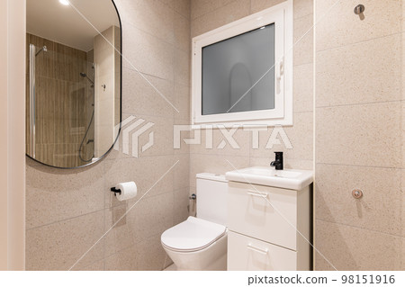 Part of bathroom with toilet bowl, washbasin on table, beautiful oval mirror on wall lined with beige marble tiles, in which you can see reflection of shower area with metal fittings. Part of bathroom with toilet bowl, washbasin on table, beautiful oval mirror on wall lined with beige marble tiles, in which you can see reflection of shower area with metal fittings. 98151916