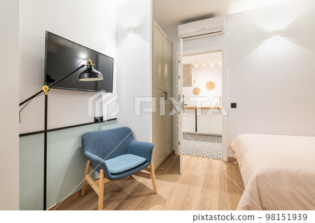 Small cozy room with bed covered with beige blanket. Comfortable blue armchair in which you can read by light of night lamp. Large TV on wall opposite bed for watching interesting movies. 98151939