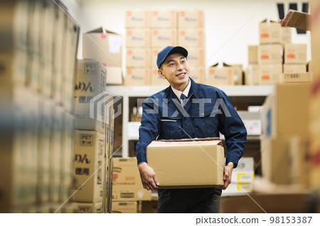 A man in work clothes carrying a cardboard box in a distribution warehouse 98153387