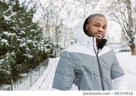 Young African man in casual white and gray clothes, sport jacket with hood . Winter season street portrait in park with snow. Lifestyle, city life concept Young African man in casual white and gray clothes, sport jacket with hood . Winter season street portrait in park with snow. Lifestyle, city life concept 98154016