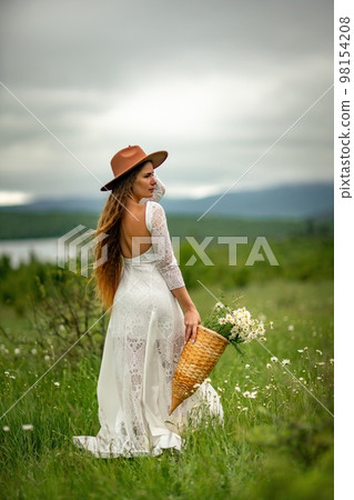A middle-aged woman in a white dress and brown hat stands on a green field and holds a basket in her hands with a large bouquet of daisies. In the background there are mountains and a lake. 98154208