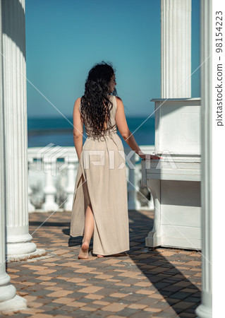 Sea woman rest. A woman with long curly hair in a beige dress stands with her back and looks at the sea and the coast from a balcony with balusters. Tourist trip to the sea. Sea woman rest. A woman with long curly hair in a beige dress stands with her back and looks at the sea and the coast from a balcony with balusters. Tourist trip to the sea. 98154223